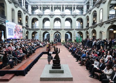 En la fotografía se observa un evento oficial en el Palacio Nacional de la Cultura, Guatemala. El salón principal muestra arquitectura colonial con arcos y balcones en dos niveles. Dos grupos grandes de personas están sentadas frente a un podio central, con una escultura en primer plano. A la izquierda hay una pantalla grande con un código QR y una imagen promocional. Al fondo, fotógrafos y camarógrafos documentan el acto.