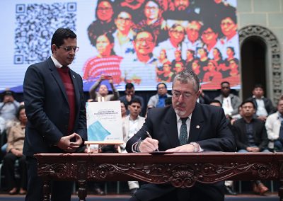 En la fotografía se observa en primer plano al Presidente Bernardo Arévalo firmando un documento sobre una mesa tallada de madera en el Palacio Nacional de la Cultura, Guatemala. Frente a ella hay un atril con una carpeta que muestra el título “Protección de Personas Defensoras de Derechos Humanos”. Al fondo, se observa una pantalla grande con una imagen colorida y varias personas sentadas en filas.