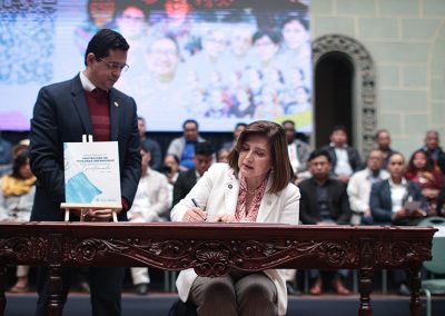 En la fotografía se observa en primer plano a la Vicepresidenta Karin Herrera firmando un documento sobre una mesa tallada de madera en el Palacio Nacional de la Cultura, Guatemala. Frente a ella hay un atril con una carpeta que muestra el título “Protección de Personas Defensoras de Derechos Humanos”. Al fondo, se observa una pantalla grande con una imagen colorida y varias personas sentadas en filas.