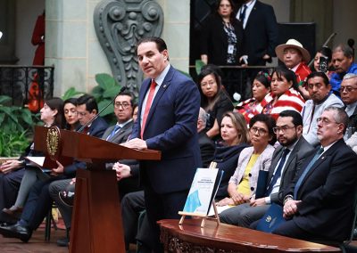 En la imagen se observa al Ministro de Gobernación, Marco Antonio Villeda, de pie detrás de un podio de madera con el escudo nacional, ofreciendo un discurso en un salón del Palacio Nacional de la Cultura. Al fondo, varias personas están sentadas en filas, algunas con vestimenta formal y otras convestimenta maya en donde predominan los colores rojo y blanco. La decoración incluye plantas verdes y detalles arquitectónicos tallados en piedra.