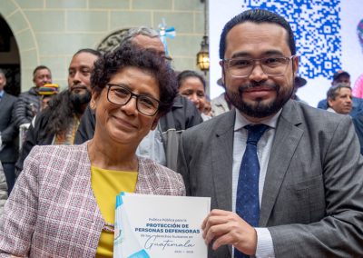 Makrina Gudiel, representante de la Familia Gudiel Álvarez, y Elvyn Díaz, Director Ejecutivo de la COPADEH, sostienen un documento titulado “Política Pública para la Protección de Personas Defensoras de los Derechos Humanos en Guatemala 2025-2035”. Ambos están de pie en un espacio público con personas y una pantalla grande en el fondo que muestra un código QR y parte de una imagen promocional.