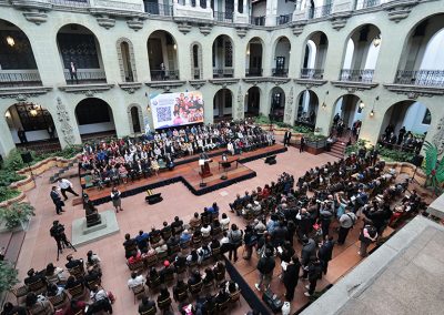 Fotografía tomada desde lo alto, se observa un evento oficial en el Palacio Nacional de la Cultura, Guatemala. El salón principal muestra arquitectura colonial con arcos y balcones en dos niveles. Dos grupos grandes de personas están sentadas frente a un podio central, a la izquierda una escultura. Detras de las personas hay una pantalla grande con un código QR y una imagen promocional. A los costados, fotógrafos y camarógrafos documentan el acto.