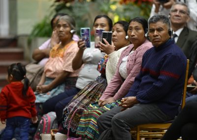 En la imagen se observa a integrantes del público, principalmente mujeres y hombres indígenas sentados en filas, quienes portan dispositivos de interpretación simultánea y graban el acto con sus teléfonos móviles.