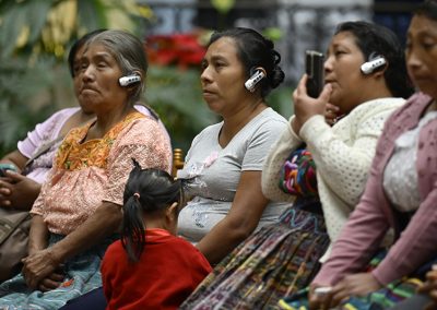 En la imagen se observa a integrantes del público, principalmente mujeres y hombres indígenas sentados en filas, quienes portan dispositivos de interpretación simultánea y graban el acto con sus teléfonos móviles durante el acto de disculpas públicas del Estado de Guatemala por el caso Pérez Lucas Vs. Guatemala, realizado en el Palacio Nacional de la Cultura.