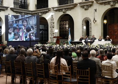 En la imagen se observa el acto de disculpa pública del Estado de Guatemala realizado en el Palacio Nacional de la Cultura, al fondo la mesa principal y en primer plano el público asistente sentado frente a ellos y una pantalla que proyecta imágenes relacionadas con el caso Pérez Lucas Vs. Guatemala.
